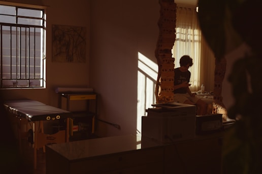 A dimly lit room with a person sitting by a window, partially illuminated by soft, warm light. The room has a massage table, some equipment, and artwork on the wall. Shadows cast interesting patterns, adding depth to the serene space.