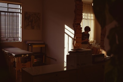 A dimly lit room with a person sitting by a window, partially illuminated by soft, warm light. The room has a massage table, some equipment, and artwork on the wall. Shadows cast interesting patterns, adding depth to the serene space.