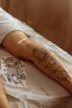A person's forearm is resting on a surface with a freshly drawn tattoo outline visible. The tattoo design appears to be inspired by a fantasy character, incorporating a figure with ornate armor and decorative elements. A paper with a similar sketch is lying beside the arm, possibly used as a reference for the tattoo. The scene is warmly lit, creating a cozy ambiance.