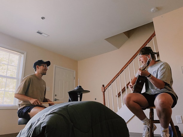 Two people are engaged in a musical session indoors. One person, seated on the left, is playing an electronic drum kit while the other, seated on the right, is playing an acoustic guitar and singing. The room includes a window, a closed door, and a staircase leading upwards. Both individuals are casually dressed and appear to be enjoying the moment.