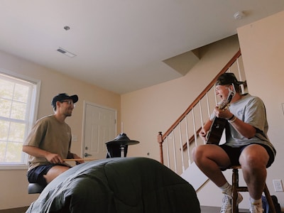 Two people are engaged in a musical session indoors. One person, seated on the left, is playing an electronic drum kit while the other, seated on the right, is playing an acoustic guitar and singing. The room includes a window, a closed door, and a staircase leading upwards. Both individuals are casually dressed and appear to be enjoying the moment.