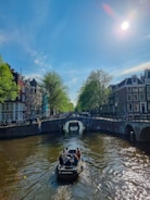 A boat with several people is traveling along a canal, surrounded by historic buildings and trees on either side. The sky is clear with the sun shining brightly, casting reflections on the water. A bridge crosses the canal in the background, with people walking and biking.