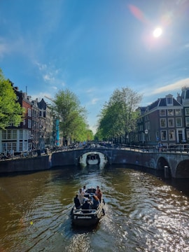 A boat with several people is traveling along a canal, surrounded by historic buildings and trees on either side. The sky is clear with the sun shining brightly, casting reflections on the water. A bridge crosses the canal in the background, with people walking and biking.
