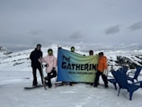 A group of people dressed in winter gear hold a banner that reads 'The Gathering Youth Performance Co.' They are standing on a snowy landscape with mountains in the background. Two snowboards are visible, and there are a few blue chairs on the right.