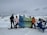 A group of people dressed in winter gear hold a banner that reads 'The Gathering Youth Performance Co.' They are standing on a snowy landscape with mountains in the background. Two snowboards are visible, and there are a few blue chairs on the right.