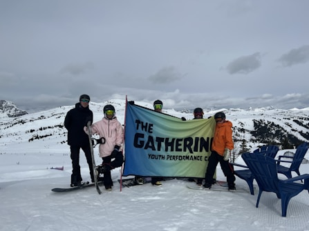 A group of people dressed in winter gear hold a banner that reads 'The Gathering Youth Performance Co.' They are standing on a snowy landscape with mountains in the background. Two snowboards are visible, and there are a few blue chairs on the right.
