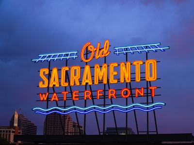 A neon sign displaying the words 'Old Sacramento Waterfront' in a vibrant combination of blue, yellow, red, and orange lights. The sky in the background is a deep blue-purple. Modern buildings are partially visible behind the sign.