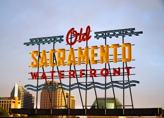 A colorful neon sign stating 'Old Sacramento Waterfront' positioned against a backdrop of modern buildings under a clear sky. The words 'Old' and 'Waterfront' are in red, while 'Sacramento' is in yellow, with green wave-like designs and borders. Skyscrapers and office buildings are visible in the background.