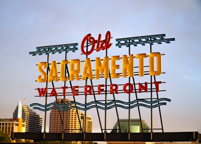 A colorful neon sign stating 'Old Sacramento Waterfront' positioned against a backdrop of modern buildings under a clear sky. The words 'Old' and 'Waterfront' are in red, while 'Sacramento' is in yellow, with green wave-like designs and borders. Skyscrapers and office buildings are visible in the background.