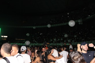 A crowded indoor venue filled with people standing and socializing. Large white balloons float above the crowd. The setting appears to be lively and festive with dim lighting accentuated by a green light source on one side.