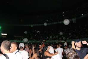 A crowded indoor venue filled with people standing and socializing. Large white balloons float above the crowd. The setting appears to be lively and festive with dim lighting accentuated by a green light source on one side.