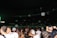 A crowded indoor venue filled with people standing and socializing. Large white balloons float above the crowd. The setting appears to be lively and festive with dim lighting accentuated by a green light source on one side.