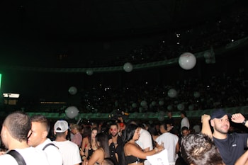 A crowded indoor venue filled with people standing and socializing. Large white balloons float above the crowd. The setting appears to be lively and festive with dim lighting accentuated by a green light source on one side.