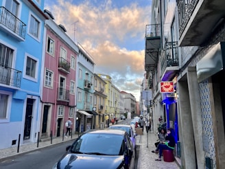 A narrow street lined with colorful buildings, including shades of blue, pink, and yellow. Several people are walking and others are seated outside a bar with a neon sign that reads 'Bar Piripi'. Parked cars are along the roadside. The sky is partly cloudy with a warm, golden hue from the setting sun.