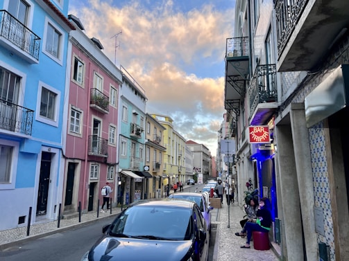 A narrow street lined with colorful buildings, including shades of blue, pink, and yellow. Several people are walking and others are seated outside a bar with a neon sign that reads 'Bar Piripi'. Parked cars are along the roadside. The sky is partly cloudy with a warm, golden hue from the setting sun.