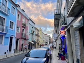 A narrow street lined with colorful buildings, including shades of blue, pink, and yellow. Several people are walking and others are seated outside a bar with a neon sign that reads 'Bar Piripi'. Parked cars are along the roadside. The sky is partly cloudy with a warm, golden hue from the setting sun.