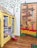 A whimsical indoor scene features a bookshelf styled as a yellow house with a roof made of books. A pair of striped stockings with red shoes emerges from underneath, reminiscent of a character from 'The Wizard of Oz.' Adjacent, a door is creatively decorated to resemble a colorful path with directional signs pointing to 'Emerald City,' 'Munchkin Land,' and other locations from the same classic story. The scene is set against a backdrop of painted green trees on the wall.