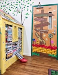 A whimsical indoor scene features a bookshelf styled as a yellow house with a roof made of books. A pair of striped stockings with red shoes emerges from underneath, reminiscent of a character from 'The Wizard of Oz.' Adjacent, a door is creatively decorated to resemble a colorful path with directional signs pointing to 'Emerald City,' 'Munchkin Land,' and other locations from the same classic story. The scene is set against a backdrop of painted green trees on the wall.