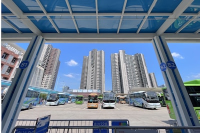A bus terminal located in an urban environment, surrounded by tall residential buildings. Several buses of various colors, including white, green, and orange, are parked in the terminal. The sky is clear with a few clouds, and there are signs with labels like B1 and B2.