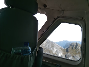 The interior perspective of an aircraft cabin is visible, featuring a close-up of a passenger's seat with a safety briefing card and bottled water in the seat pocket. Through the window, rugged mountains and a clear sky are visible.