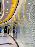 A brightly lit luxury store interior, featuring sleek, curving architecture and glossy surfaces. The walls have a reflective quality with patterned designs, and there are prominent brand logos displayed on the exterior. The color palette consists of warm gold tones complemented by neutral grays and blacks.