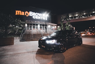 A sleek black car is parked in front of an illuminated building with signage that reads 'Mary Brown Centre.' The building features steps and a box office area. The scene is set at night with minimal street lighting and trees visible around the building.