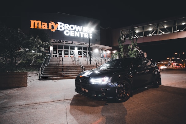 A sleek black car is parked in front of an illuminated building with signage that reads 'Mary Brown Centre.' The building features steps and a box office area. The scene is set at night with minimal street lighting and trees visible around the building.