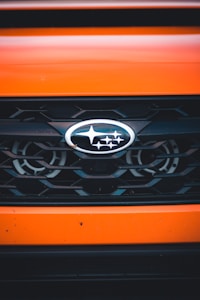 A close-up view of a car grille featuring the Subaru logo, which consists of several silver stars on a blue oval background. The grille has a black mesh design, and the car's exterior is painted in a bright orange color.