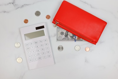 A flatlay composition featuring a white calculator positioned on a white marble surface, surrounded by various coins. Next to the calculator is a red wallet with a visible 100-dollar bill partially tucked under it. The combination of the wallet, money, and calculator suggests themes of finance or budgeting.