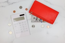 A flatlay composition featuring a white calculator positioned on a white marble surface, surrounded by various coins. Next to the calculator is a red wallet with a visible 100-dollar bill partially tucked under it. The combination of the wallet, money, and calculator suggests themes of finance or budgeting.