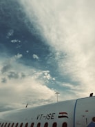 The image captures a segment of an airplane with a visible registration number and the Indian flag on its body against a backdrop of a cloudy sky. The airplane's fuselage curves toward the right edge of the photo, and the sky transitions from scattered, fluffy clouds to a clearer atmosphere.