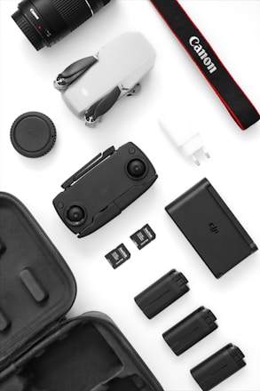 A collection of photography and drone equipment neatly arranged on a white surface. There is a camera lens, a drone, a camera strap with the Canon logo, several batteries, a battery charger, and memory cards. The black and white color scheme gives it a professional and sleek appearance.