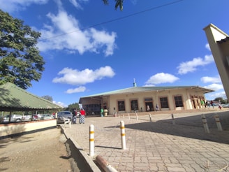 A medium-sized building with a green roof is situated in an open area under a bright blue sky with scattered clouds. Several people are walking near the entrance, and a few cars are parked nearby. There are trees with green leaves providing some shade, and white and yellow bollards are lined up along a paved area in front of the building.
