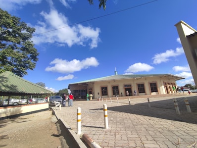 A medium-sized building with a green roof is situated in an open area under a bright blue sky with scattered clouds. Several people are walking near the entrance, and a few cars are parked nearby. There are trees with green leaves providing some shade, and white and yellow bollards are lined up along a paved area in front of the building.