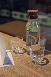 A glass bottle filled with detox water infused with dried cucumber and ginger slices, set against a soft beige background
