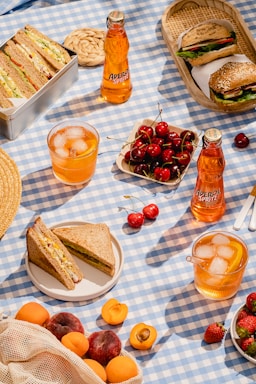 A picnic setup on a blue and white checkered cloth featuring a variety of foods and drinks. There are several sandwich halves, one in a metal box and another on a plate. A wicker basket holds a round sandwich with sesame seeds. Two bottles and two glasses of Aperol Spritz are visible, along with a bowl of fresh cherries and some scattered strawberries and apricots.