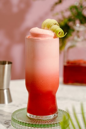 A tall glass of a frothy, layered pink and red beverage garnished with a curled lemon peel. The drink rests on a green glass coaster, with a blurred background featuring a metallic shaker and some foliage.