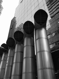 A black and white photograph featuring a row of five large, industrial metal pipes standing vertically in front of a modern glass building. The pipes have an imposing presence and a sleek design, with vent-like openings at the top. The building facade reflects some of the surrounding urban architecture, adding depth to the scene.