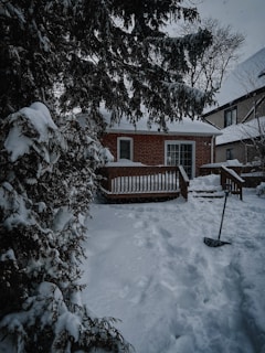 A quaint brick house with a snow-covered roof and a small wooden deck in the foreground. The yard and surrounding trees are blanketed in fresh snow, and a shovel is visible near the deck, indicating recent or ongoing snow clearing efforts. Large evergreen trees frame the scene, adding a touch of nature and providing contrast against the white snow.