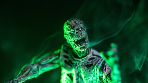 A skeletal figure with exposed bones and decaying skin, illuminated by an eerie green light. Mist or smoke partially obscures the figure, creating a haunting atmosphere.