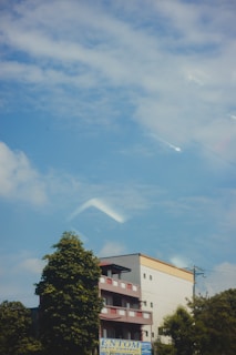 A multi-story building with a red and white exterior stands against a background of a clear blue sky with scattered clouds. There are trees partially obscuring the building, adding greenery to the urban environment. A sign at the bottom indicates a pest control business.