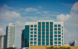 A cluster of modern high-rise buildings with large glass windows against a blue sky dotted with white clouds. In the foreground, there is a prominent building with white framing and teal glass, flanked by other skyscrapers.