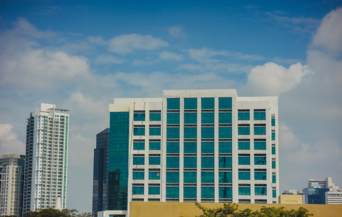 A cluster of modern high-rise buildings with large glass windows against a blue sky dotted with white clouds. In the foreground, there is a prominent building with white framing and teal glass, flanked by other skyscrapers.