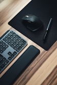 A sleek, modern workspace setup featuring a black computer keyboard with a wrist rest, a black ergonomic wireless mouse on a mouse pad, and a stylus pen. The desk has a light wood finish, contrasting with the black peripherals.