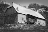 A rustic cabin stands in a mountainous rural setting, featuring a wooden and stone exterior with a metal roof. The structure is set against a backdrop of snow-dappled mountains, with sparse vegetation in the foreground, capturing a serene and rugged landscape.