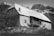 A rustic cabin stands in a mountainous rural setting, featuring a wooden and stone exterior with a metal roof. The structure is set against a backdrop of snow-dappled mountains, with sparse vegetation in the foreground, capturing a serene and rugged landscape.