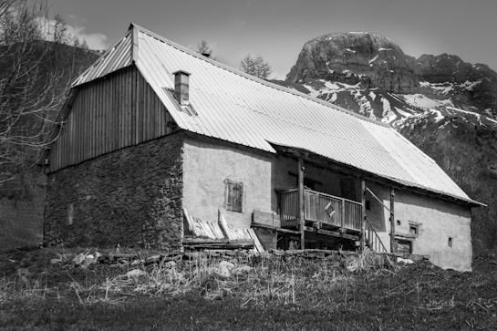 A rustic cabin stands in a mountainous rural setting, featuring a wooden and stone exterior with a metal roof. The structure is set against a backdrop of snow-dappled mountains, with sparse vegetation in the foreground, capturing a serene and rugged landscape.