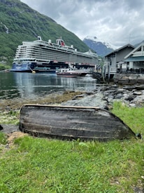 A large cruise ship is docked alongside a picturesque fjord surrounded by lush green mountains and a cloudy sky. The foreground features an overturned old wooden boat resting on grassy ground, leading to the rocky shoreline. Nearby, smaller boats are moored at a pier next to a building with a dark roof.
