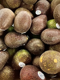 A pile of avocados with varying shades of dark green and brown, some with stickers indicating they are from Mexico.