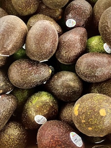 A pile of avocados with varying shades of dark green and brown, some with stickers indicating they are from Mexico.